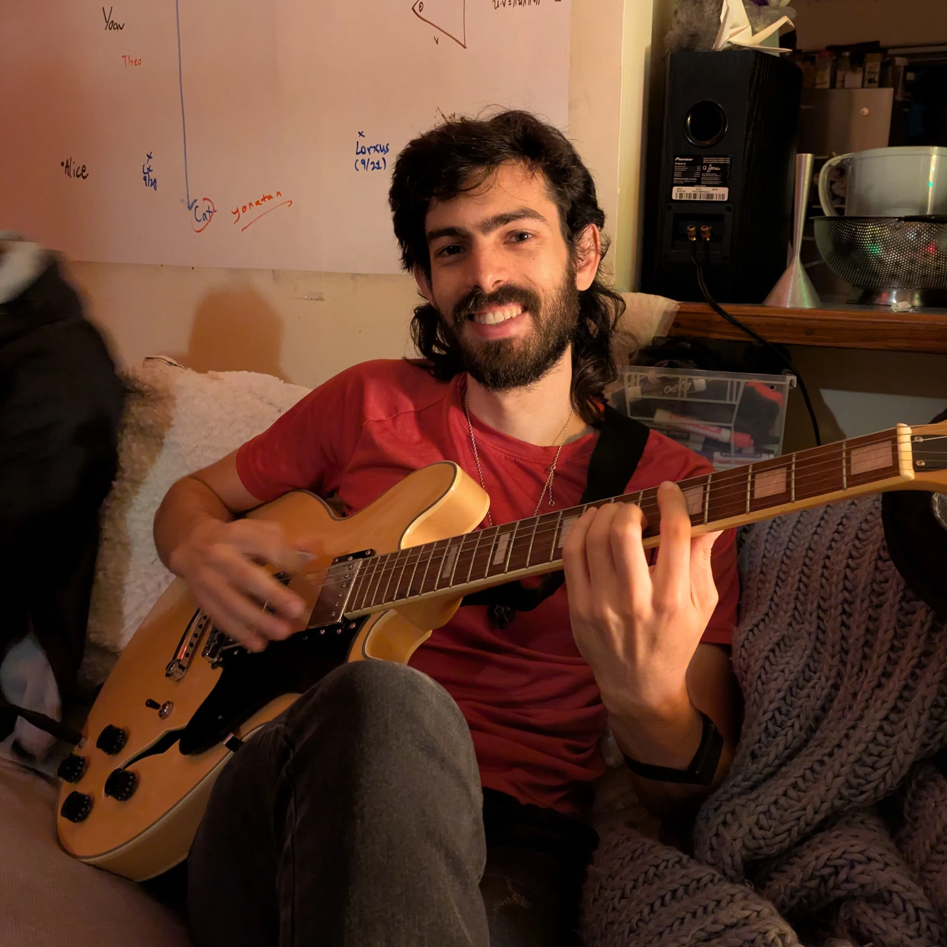 Yoav smiling on a couch, playing an electric guitar in front of a whiteboard full of doodles