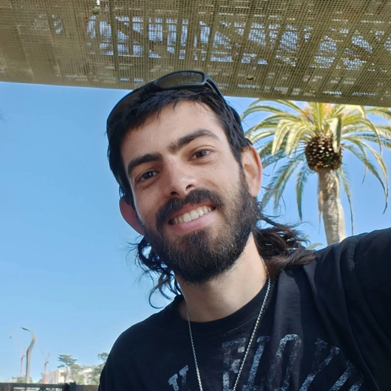 Yoav smiling in the sun, sunglasses on his head, a palm tree behind him