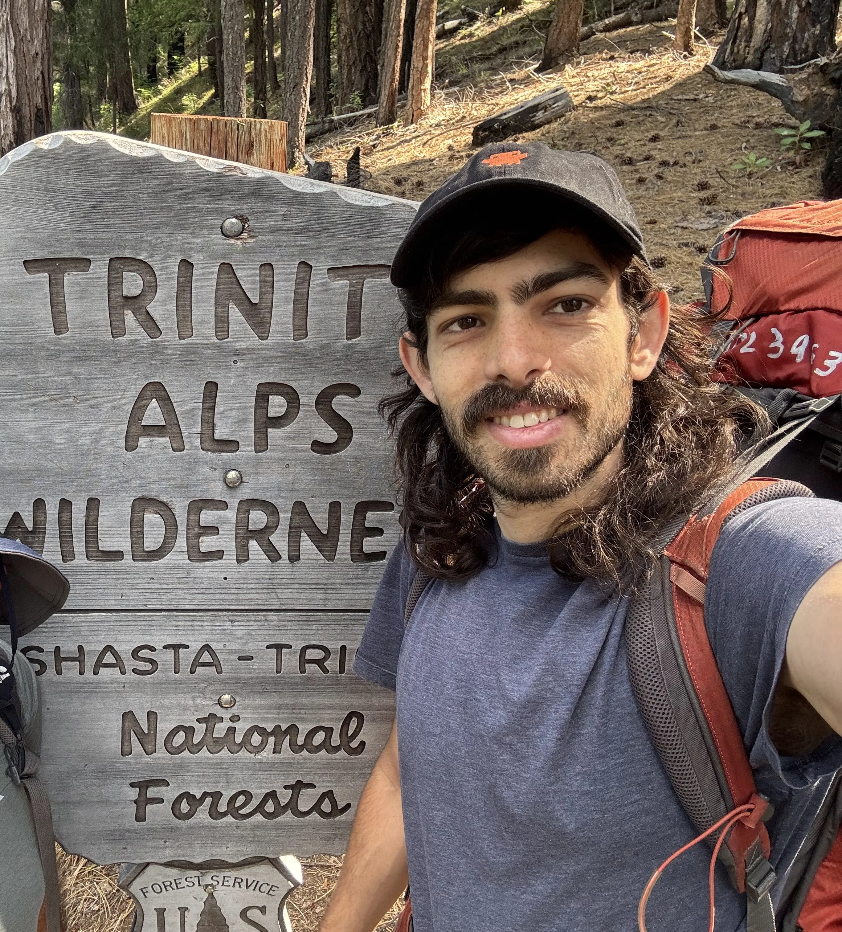 Yoav with a backpacking pack at the Trinity Alps Wilderness trailhead sign, his dad peeking in from the left